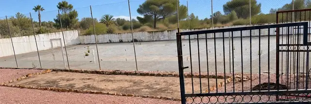 Tennisbaan vóór de renovatie met verouderde ondergrond en onkruid; auto verwijderd en achtergrond digitaal vervangen door mediterraan groen